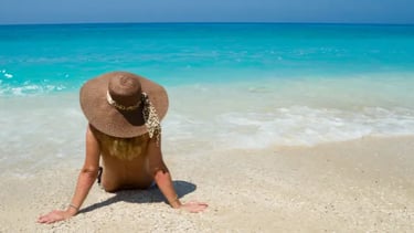 A woman wearing a sun hat sitting on a tropical beach overlooking turquoise ocean water.