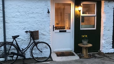White door and shop front, an old bicycle leaning against the wall. The lights are on inside.