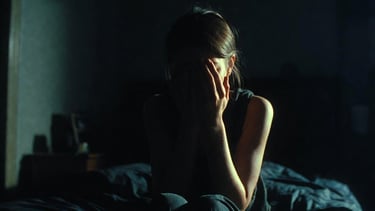 A depressed woman sitting on a bed in a dark room covering her face with her hands.
