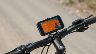 Tight shot of a phone mounted on bicycle handlebars, displaying a bright orange speed and navigation dashboard under harsh direct sunlight, dusty gravel path background.