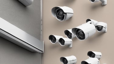 A person is standing inside a technical room or server room, working with equipment mounted on a rack. They appear focused and are adjusting or observing the setup. The equipment includes cables and a camera on a tripod. The scene is viewed through a square window with bolts, suggesting a secure or restricted area.