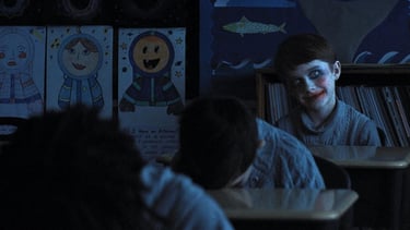 Duas crianças em uma sala de aula escura, com uma criança olhando assustadoramente para a câmera.