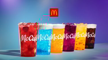 A row of colorful McCafé iced fruit drinks with foam toppings and a McDonald's logo overhead.