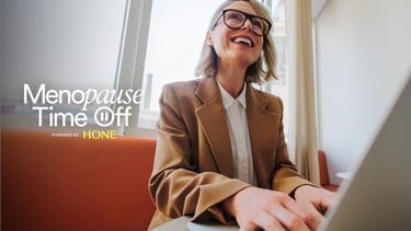 A smiling mature woman in a blazer working on a laptop, representing menopause time off support at work.