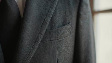 Macro close-up of hand-stitched lapel on a structured charcoal blazer, soft directional window light revealing fine textile weave, shallow depth of field.