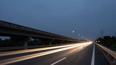 Wide panoramic view of a highway overpass at dusk, headlights streaking below in long exposure, the sky a deep blue-gray, utility poles receding into the distance on the right side, shot on 35mm with visible grain and slight lens flare from an oncoming light