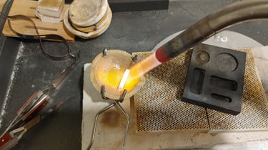 A silver jewelry-making scene showing a crucible held by tongs, heated by a torch.  