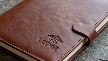 A close-up of a high-quality leather guest book with an embossed lodge logo, resting on a stone surface. Elegant, professional composition.