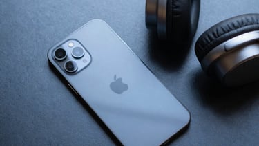 Clean, top-down photograph of modern tech hardware, including a sleek smartphone and minimalist headphones on a dark steel blue surface, North American / US.
