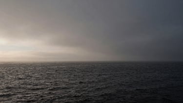 Moody 35mm landscape photography of a misty coastal horizon, deep charcoal water, soft natural light, high-contrast shadows, generous negative space.