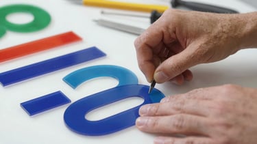Close-up, detailed shot of hands assembling acrylic letters for an illuminated sign, showing tools and materials, industrial workshop setting, crisp studio light, sharp focus on the process.