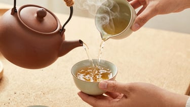 Overhead shot of hands preparing herbal tea, pouring hot water from a clay kettle into a ceramic cup, steam rising, soft morning light, warm sand tones