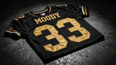 Moody low-key product photography of a vintage football jersey sponsor print, high-contrast dramatic spotlighting, deep shadows, rich saturated gold lettering on a textured black weave, dark concrete background, macro lens