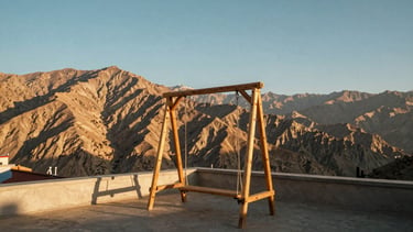 A wooden swing on a quiet rooftop terrace in Leh, panoramic view of the majestic Stok mountain range under a clear blue sky, soft golden hour light