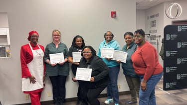 Seven women smiling while holding training certificates during a community partnership graduation event.