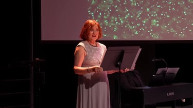 A female narrator on stage in a formal gown along with a music stand & piano.