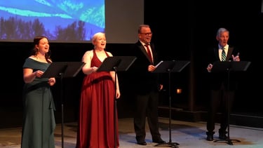 Four singers in formal attire perform a vocal concert on stage with music stands.