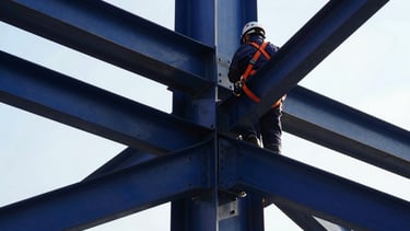 Detailed close-up of structural steel joints being bolted by a technician in full PPE, safety orange harness, high-contrast natural daylight, 35mm.