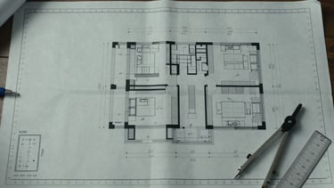 Extreme wide-angle overhead view of a detailed architectural blueprint spread flat on a white surface, precise measurement annotations and section callouts visible under cool daylight, drafting tools—a steel scale rule and compass—resting at the lower right corner, generous negative space on the left third of the frame