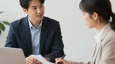 A man and a woman in business attire have a conversation. The woman takes notes as the man speaks.