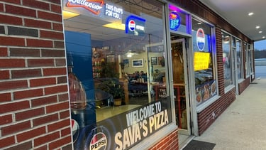 Exterior of Sava's Pizza storefront with neon beer signs and Pepsi logo on a brick building.