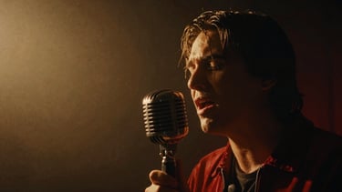 Cinematic film photography, close-up of a musician singing into a vintage microphone in a dark, hazy studio illuminated by a single warm spotlight, high-contrast shadows.