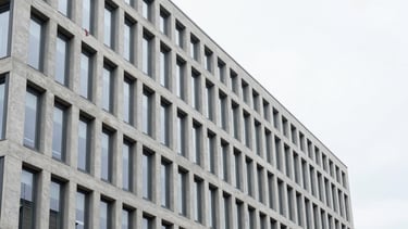 Sharp architectural photograph of a modern Swiss office building in Flaach, clean concrete and glass facade, bright overcast daylight, desaturated cool tones with a subtle red accent flag, 35mm
