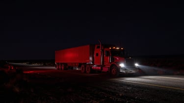 Wide panoramic Arizona desert highway at night, a heavy-duty dually truck with flatbed trailer heading towards the horizon, headlights cutting through the darkness, dramatic low-light, high-contrast, deep charcoal and red tones, 35mm.