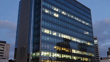 A wide photography shot of a modern corporate building in a South American / Brazilian city at dusk, with lights glowing behind large glass windows. The composition reflects expertise and reliability, using a palette of dark blue and steel blue. Soft architectural lighting creates a professional and efficient mood.