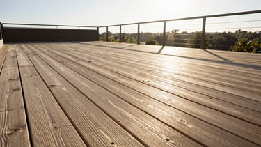 Wide environmental shot of a completed deck in afternoon sun, composite or hardwood boards running diagonally from foreground to a railing with cable detail, the boards' grain texture visible in the low-angle light, no people, no furniture, the structure speaking for itself