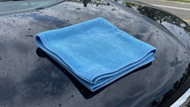 Close-up of water beautifully beading on a freshly waxed deep black car hood, electric baby blue microfiber cloth wiping the surface, high-contrast reflections.