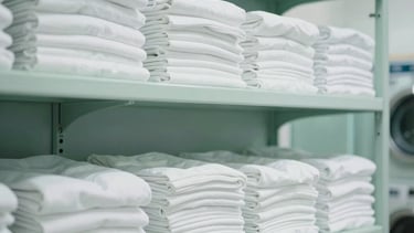 Clean and bright photography of a professional laundry facility in Porto-Vecchio, Southern Europe. Soft natural light illuminates stacks of fresh white linens on minimalist shelves. The palette features soothing shades of light blue and sage green, projecting a modern and serene atmosphere.