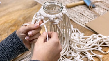 Manos haciendo nudos para un proyecto de macramé DIY alrededor de un frasco de vidrio sobre una mesa