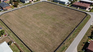 Overhead aerial view of a land parcel perimeter, lot markers clearly visible at corners, neighboring development pattern visible at the frame edge, access road running along the southern boundary, natural midday light — forensic clarity, no people