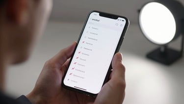 Close-up of an Android phone held in one hand, a feature list visible on screen with several items crossed out, studio light mimicking workspace brightness, over-the-shoulder framing, shallow depth of field on the device