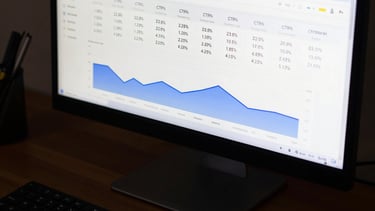 Close-up overhead view of a widescreen monitor displaying a Google Ads performance dashboard with real campaign metrics — CTR percentages, cost-per-click columns, and conversion graphs in blue and white — on a dark desk surface, even studio lighting, no people visible