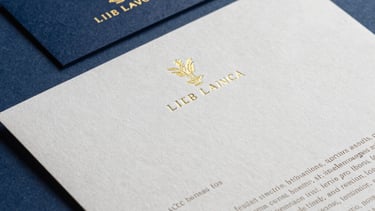 Detailed shot of printed brand stationery: business cards and letterheads with embossed gold-toned foil. Elegant, Latin American / Hispanic luxury style, moody shadows, deep blue background.