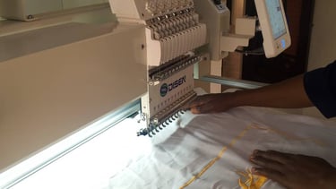 Professional multi-needle embroidery machine stitching yellow thread onto white fabric with a touchscreen control panel.