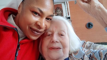 A smiling caregiver and an elderly woman holding a bed trapeze handle during home elder care.