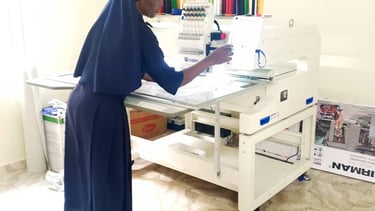 A nun in blue habit operating a white professional multi-needle embroidery machine in a workshop.