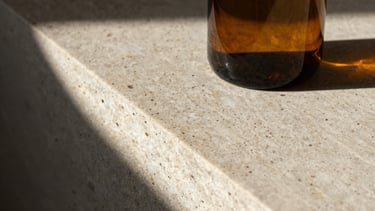 Tactile close-up of raw travertine stone texture and a heavy amber glass bottle, sharp afternoon shadows, high contrast, 35mm.