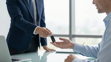 A high-resolution photography shot in a bright North American office. A professional person in a light blue shirt hands a set of car keys to a smiling client across a clean glass desk. The atmosphere is transparent and secure. Soft morning light streams through the window. The color palette emphasizes dark blue and off-white tones for a trustworthy financial service aesthetic.