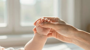 A close-up shot of a mother's hand holding her baby's tiny hand, warm sunlight filtering through a window, soft focus, 35mm editorial style.