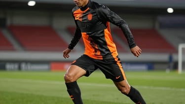 A dynamic action shot of a professional athlete training on a lush performance green soccer field at dusk. The athlete wears high-performance gear in carbon black and energy orange. Stadium lights create a professional, cinematic atmosphere. Global / Spanish-speaking context.