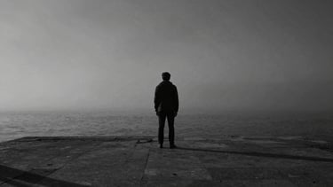 Moody cinematic still of a solitary figure standing on a misty Lisbon pier, desaturated monochrome tones, high-contrast chiaroscuro lighting, 35mm film grain.