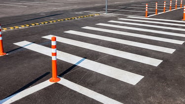 Painted white pedestrian crosswalk on asphalt with orange safety bollards in a parking lot.