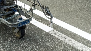 A road marking machine sprays white paint to create fresh stripes on an asphalt surface.