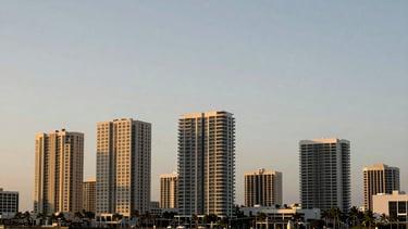 Wide panoramic Sarasota, Florida cityscape at golden hour, modern architectural buildings, quiet coastal skyline, soft natural light, muted navy and champagne gold tones, sharp focus, 35mm lens