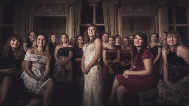 Smiling bride in white lace gown poses with her bridesmaids in formal dresses at a wedding reception.
