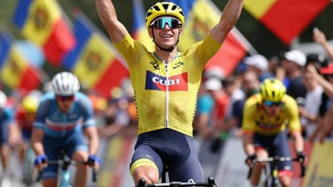 Action shot of a cyclist celebrating at the finish line, arms raised, with Moldovan flags waving gently in the background. Bright daylight, energetic atmosphere with spectators blurred in the background.